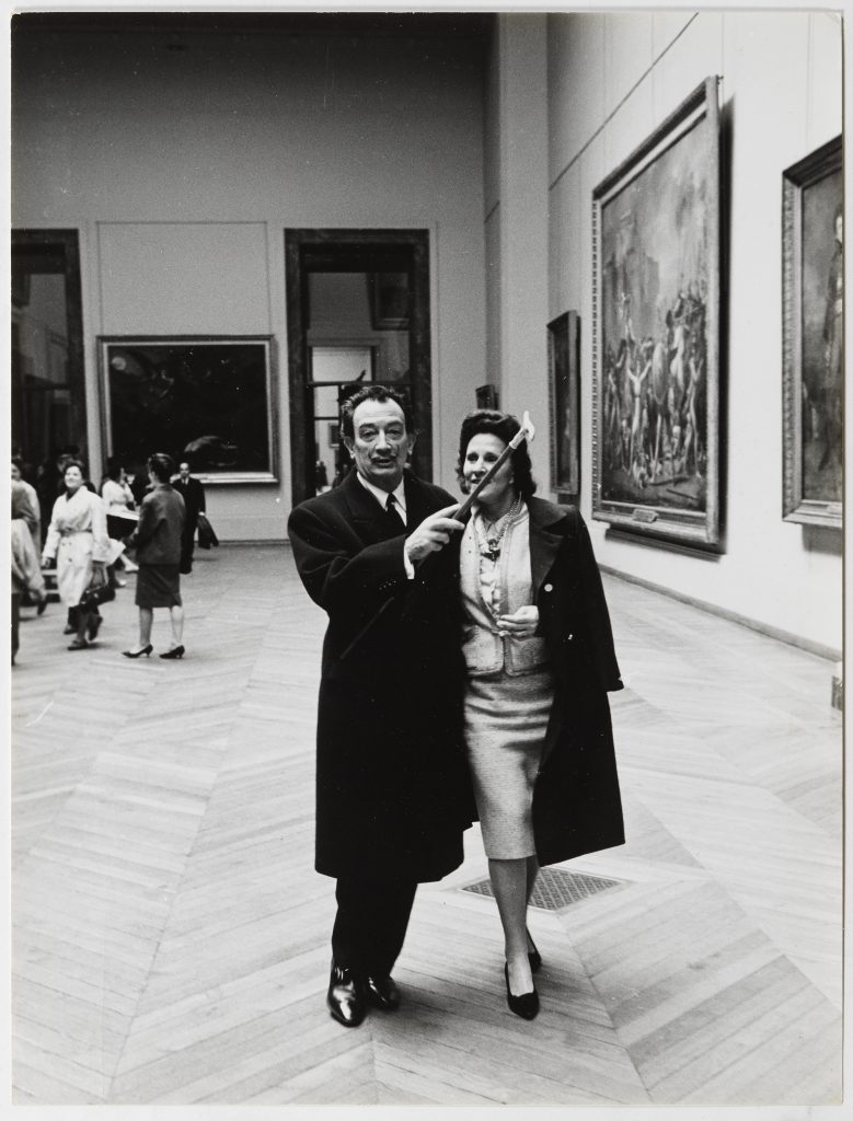 Salvador Dalí and Gala visiting the Neoclassical section of the Louvre Museum in Paris. Behind them, <em>The Intervention of the Sabine Women </em>(1799) by Jacques-Louis David, c. 1965 
Atlantic Press. Image rights of Salvador Dalí and Gala reserved. 
Fundació Gala-Salvador Dalí, Figueres, 2024